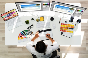 Vista superior de un diseñador gráfico trabajando en una mesa blanca con muchas pantallas, hojas y herramientas