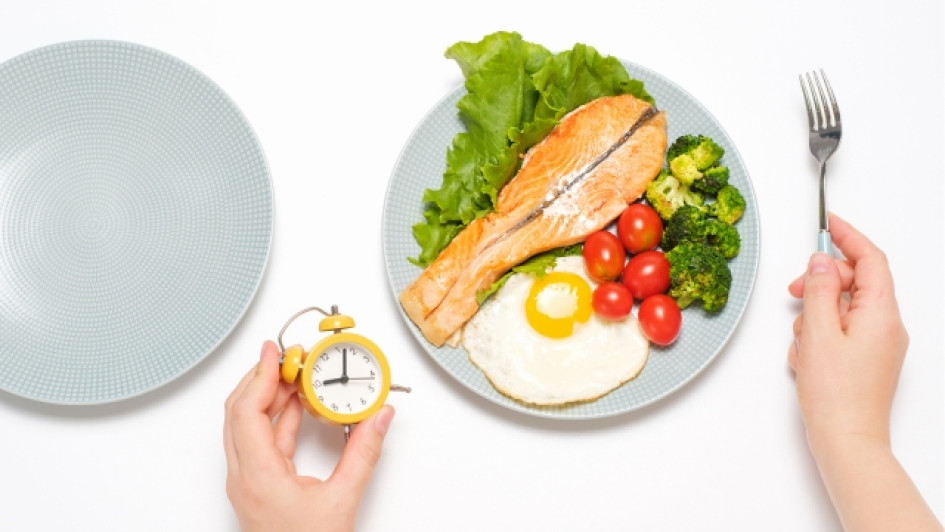 Persona sentada frente a un plato de comida sosteniendo un reloj en su mano izquierda.